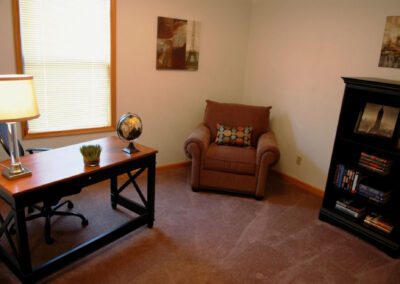 A cozy room featuring a chair, desk, lamp, and a bookshelf filled with books, creating a warm study environment.